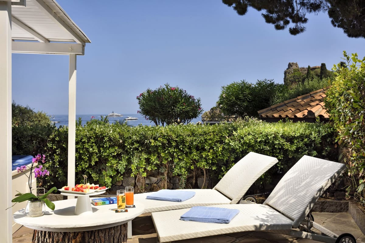 A patio with two lounge chairs, a table with a fruit platter and drinks, and a view of the sea and a castle in the background.