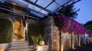 A stone staircase leading to a covered entrance with a glass roof, surrounded by lush greenery and purple flowers. - La Plage Resort (Taormina)