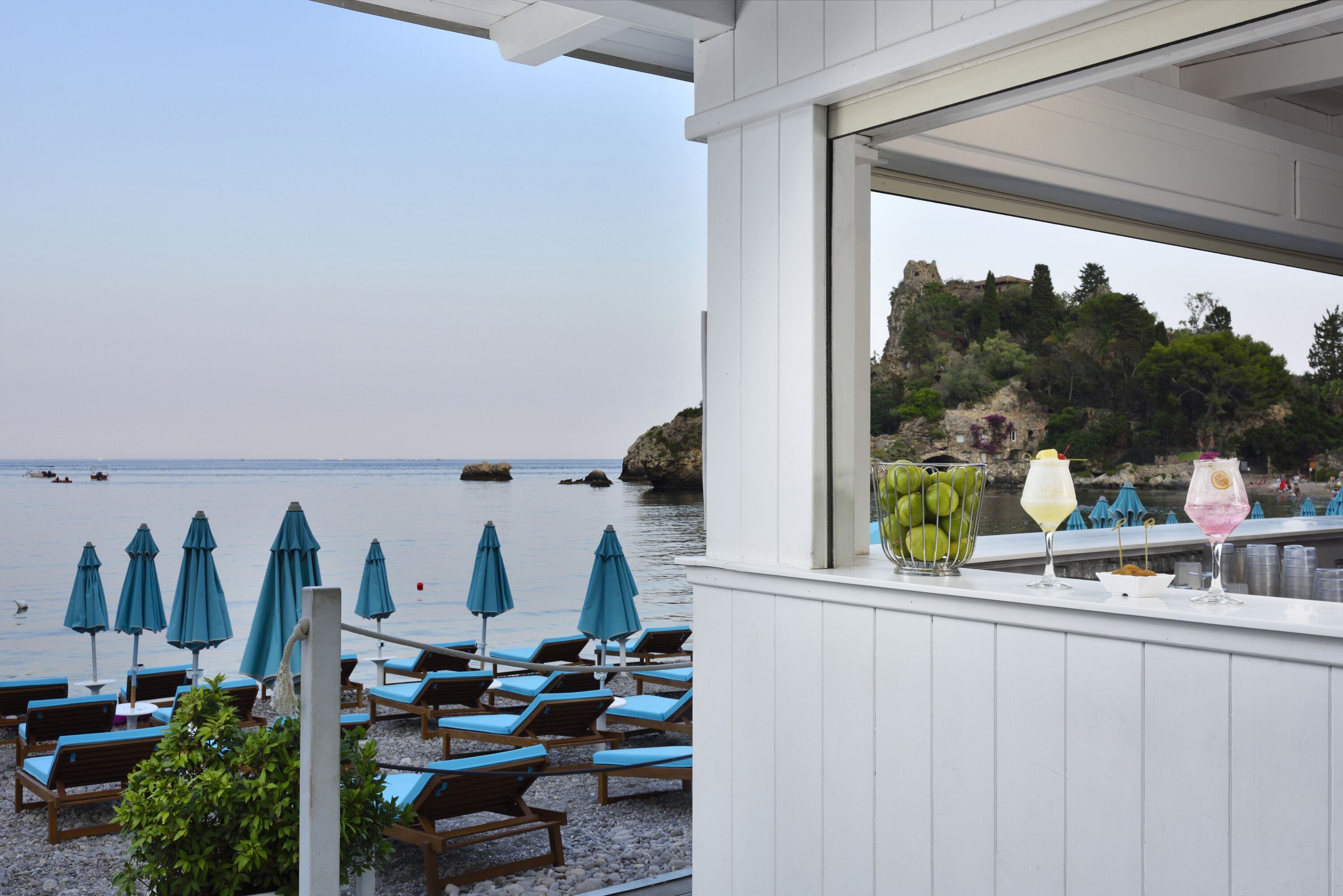 A beachside bar with blue umbrellas, lounge chairs, and a view of the sea and rocky cliffs.