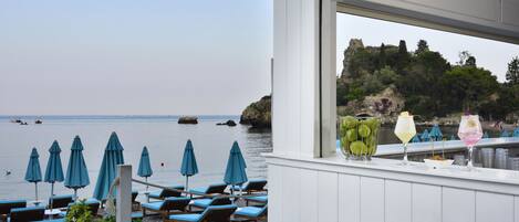 A beachside bar with blue umbrellas, lounge chairs, and a view of the sea and rocky cliffs.