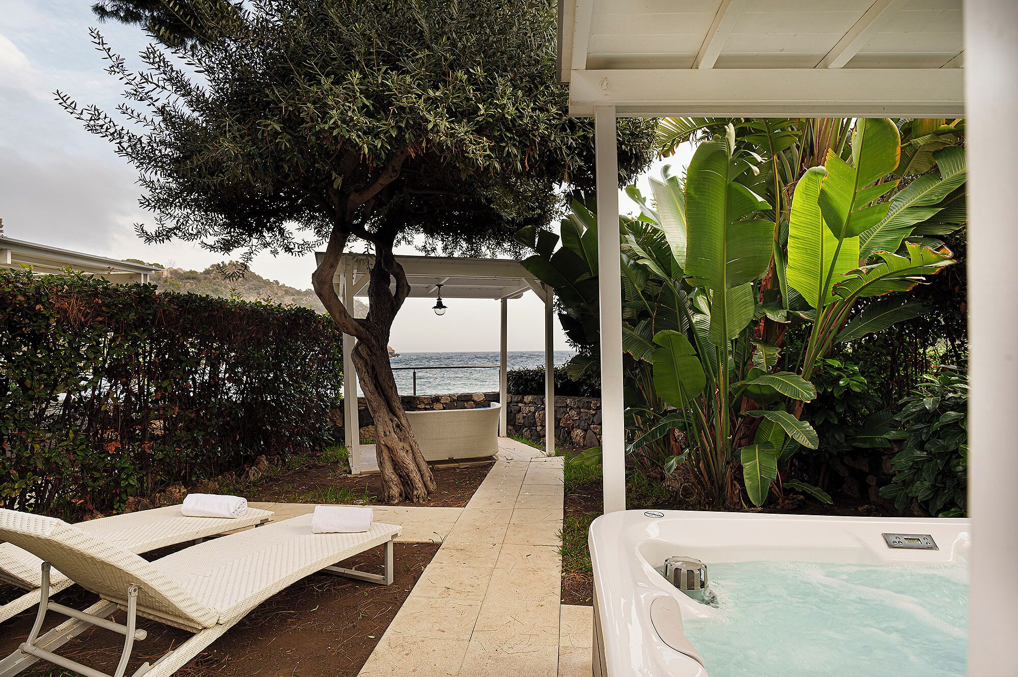 A pool area with lounge chairs, a hot tub, and a view of the sea.