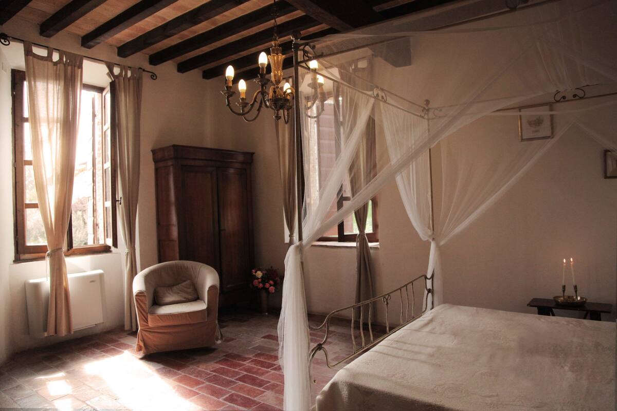 Un dormitorio con cama con dosel, una silla de madera, una mesita de noche con velas, un candelabro y una ventana con cortinas.