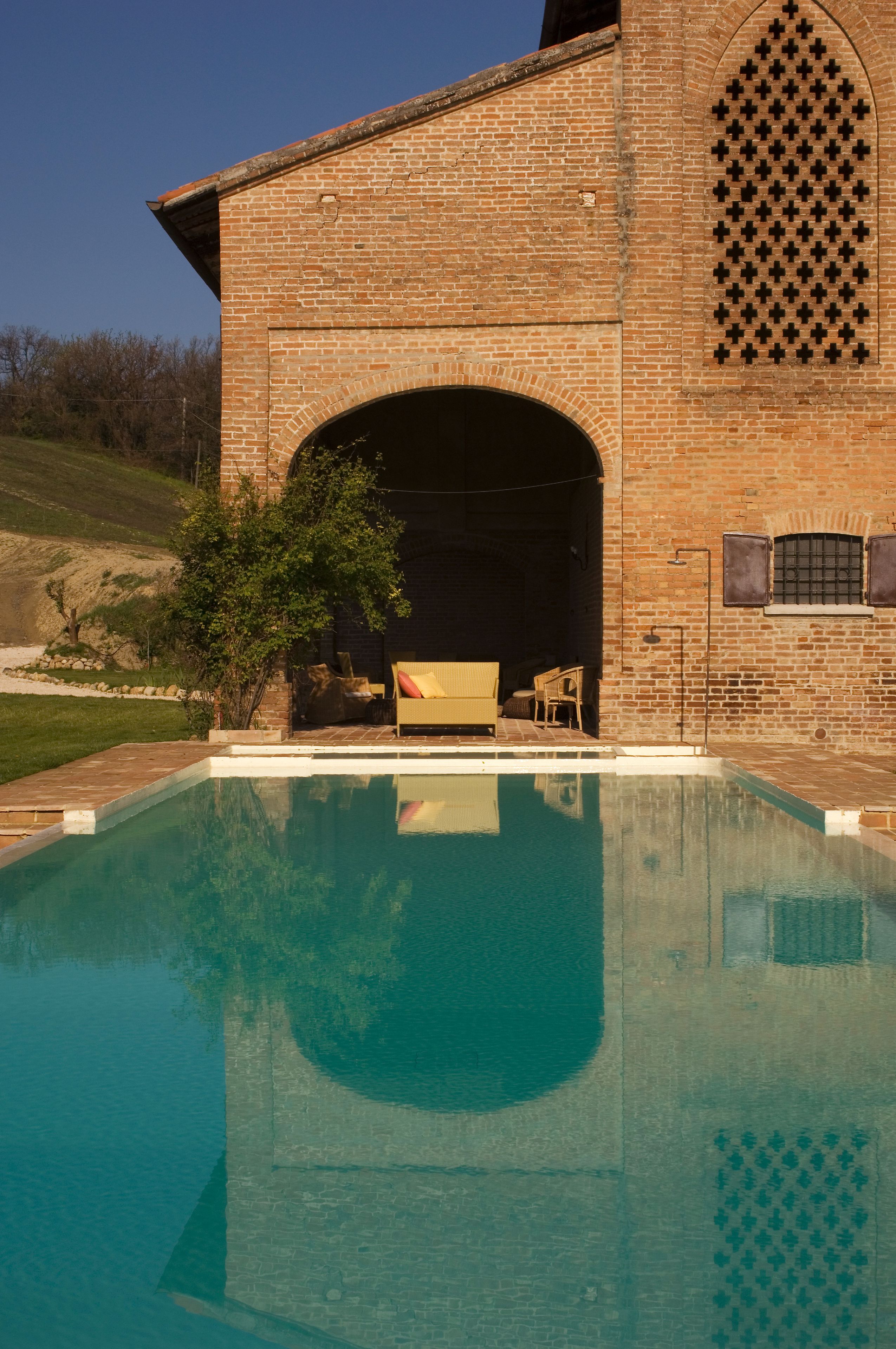 Piscina com um edifício de tijolos ao fundo, área de estar coberta e uma árvore.
