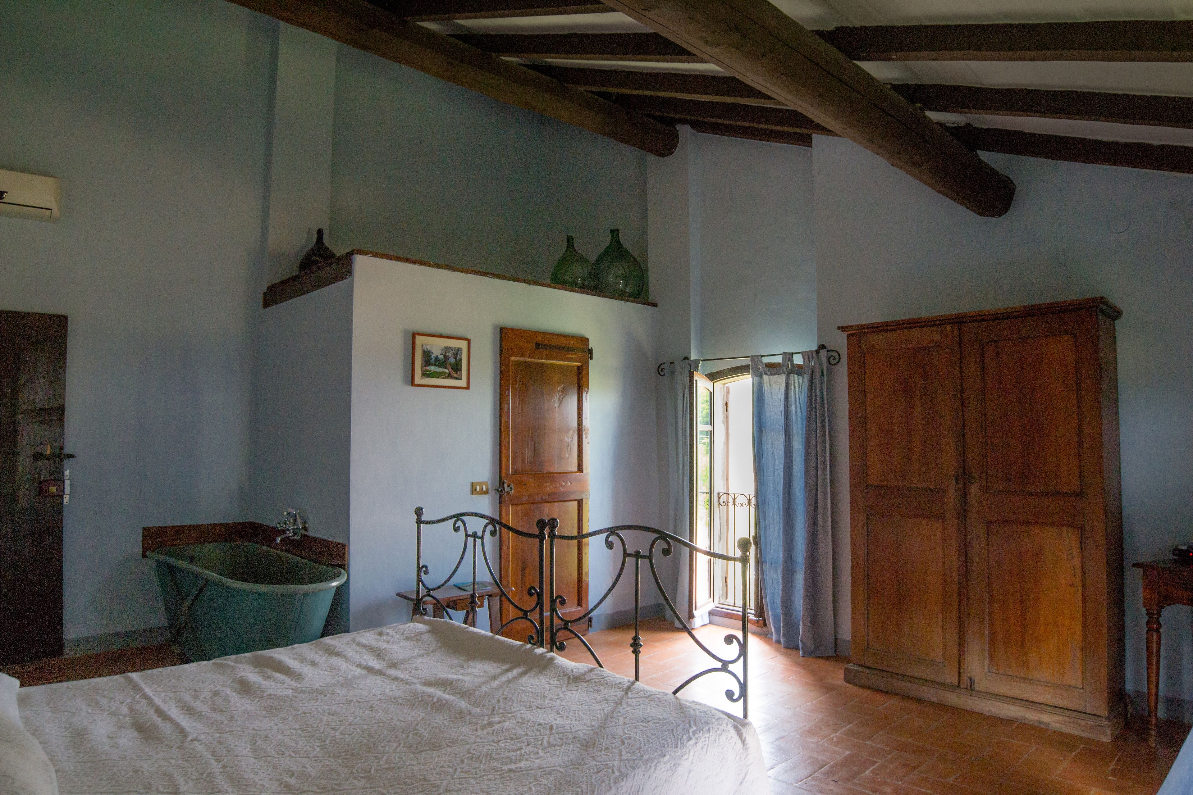 A bedroom with a bed, a wooden wardrobe, a wooden door, a green bathtub, and a window with curtains.