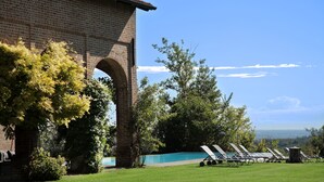 Edificio de ladrillo con entrada en arco, zona de piscina con sillas de descanso y vistas al paisaje.