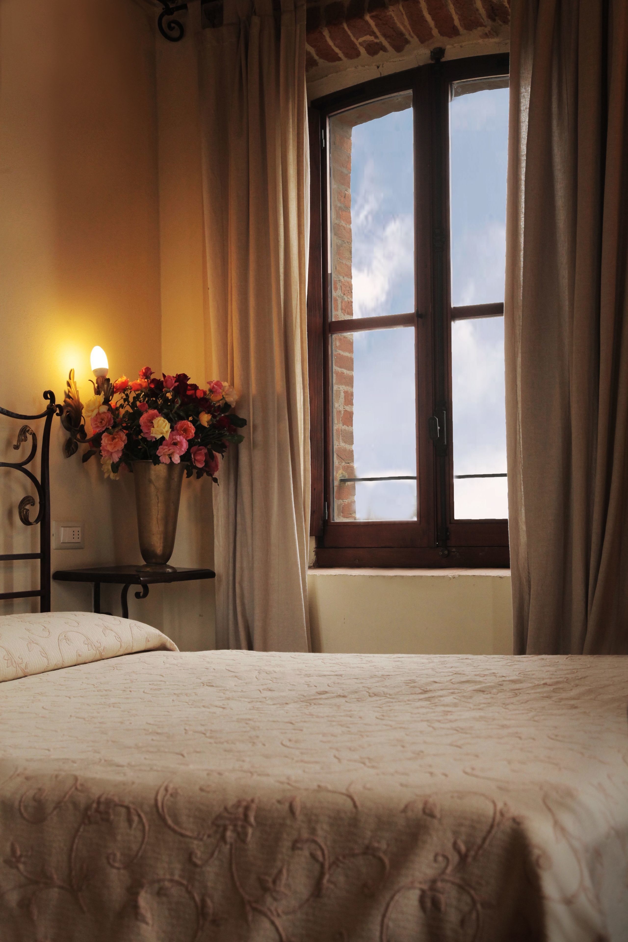 Ein Schlafzimmer mit Bett, einem Fenster mit Vorhängen, einer Wandleuchte und einer Vase mit Blumen.