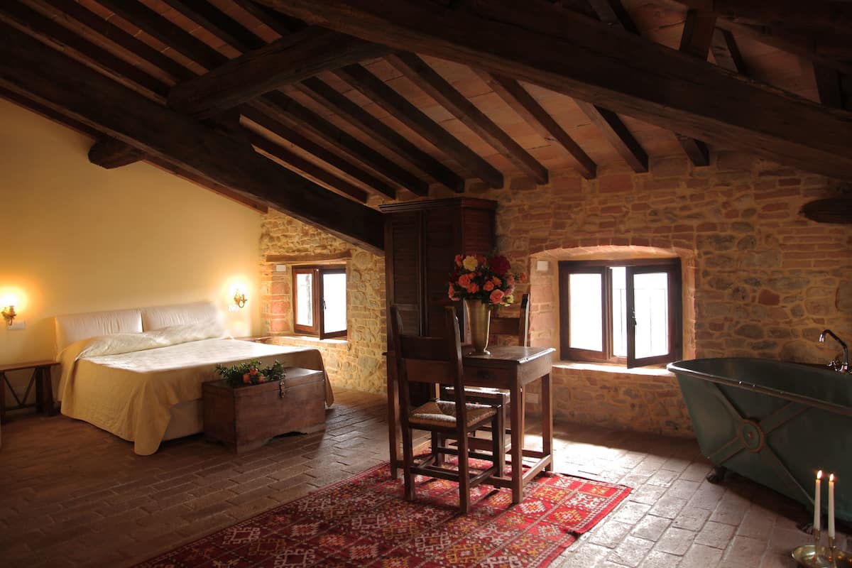Un dormitorio con cama, una silla de madera, una mesa con un florero, una pared de piedra y una bañera antigua.