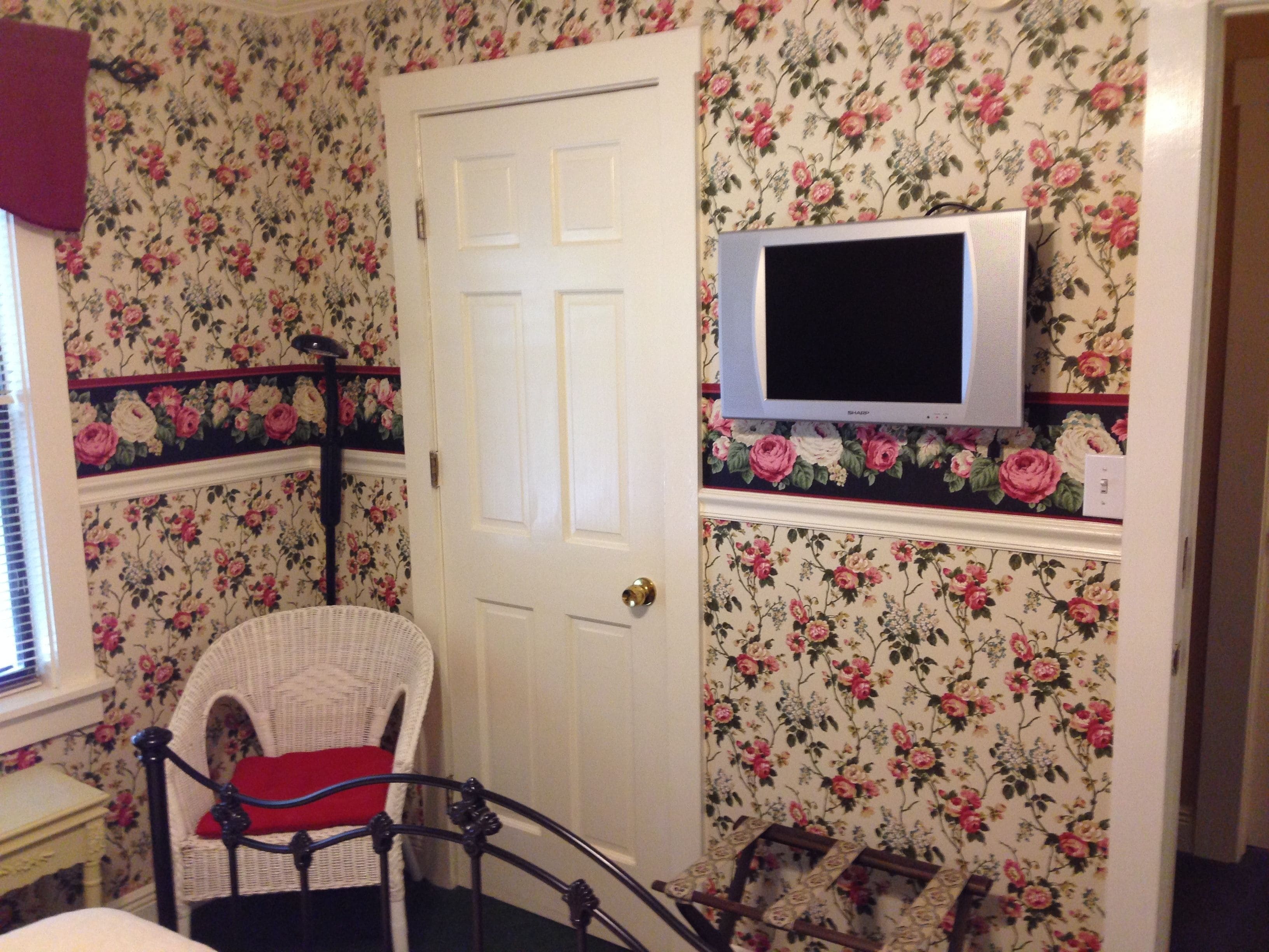 A room with floral wallpaper, a white door, a wicker chair with a red cushion, and a television mounted on the wall.