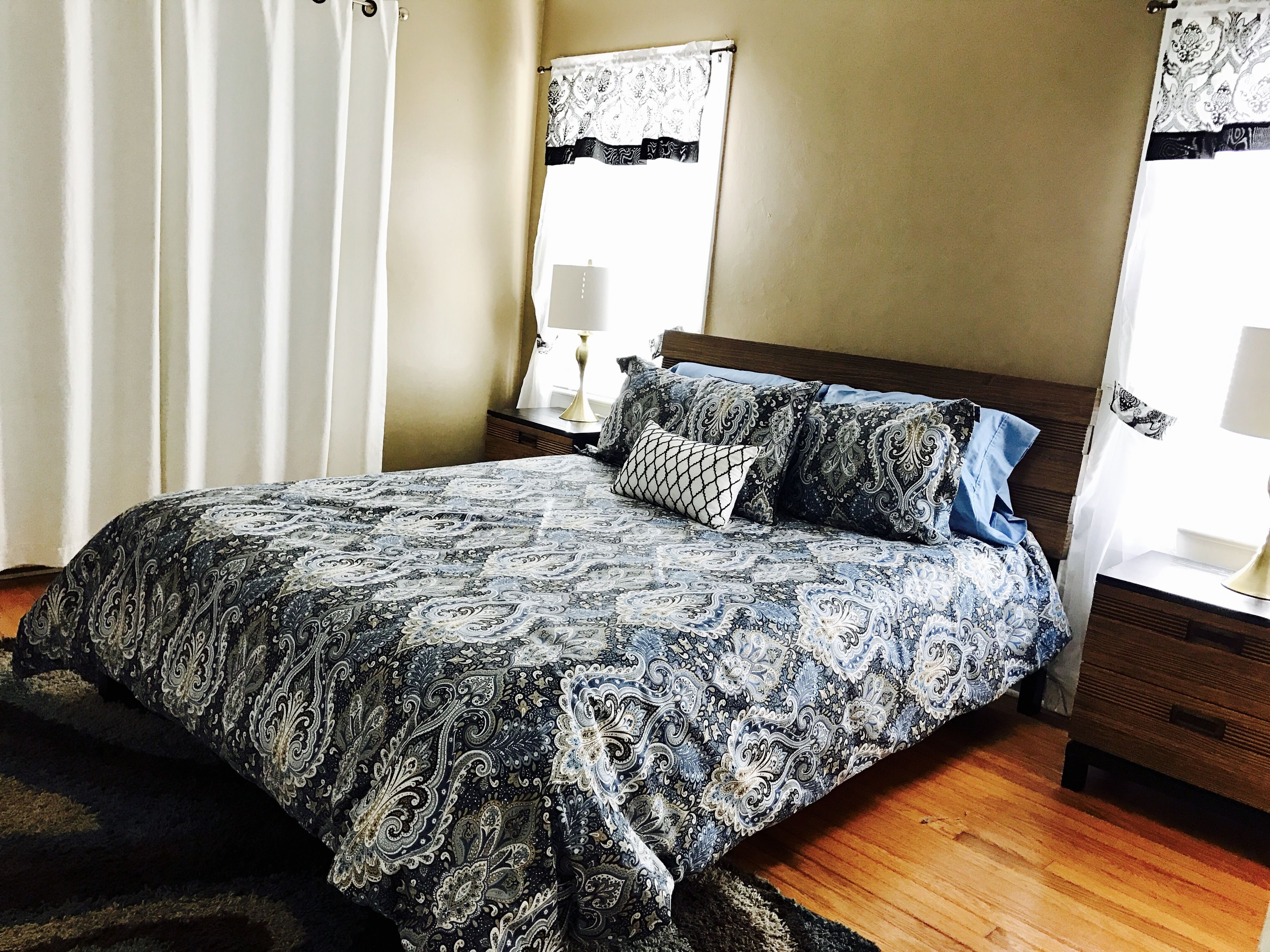 Un dormitorio con una cama grande, dos mesitas de noche, una mesilla auxiliar, una ventana con cortinas y un espejo montado en la pared.
