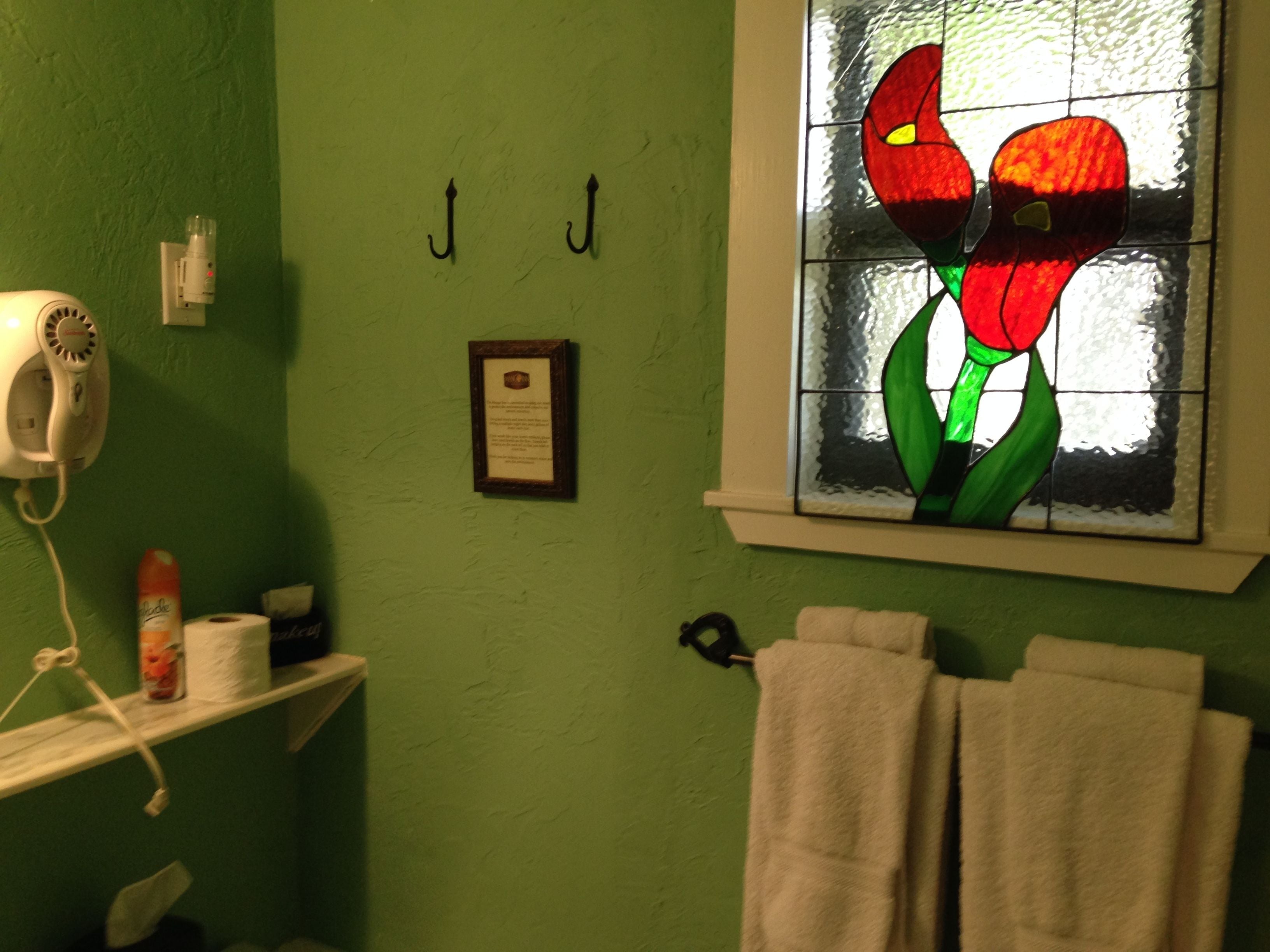 Un baño con pared verde, un perchero con dos toallas, un certificado enmarcado y una ventana con un diseño floral de vitral.
