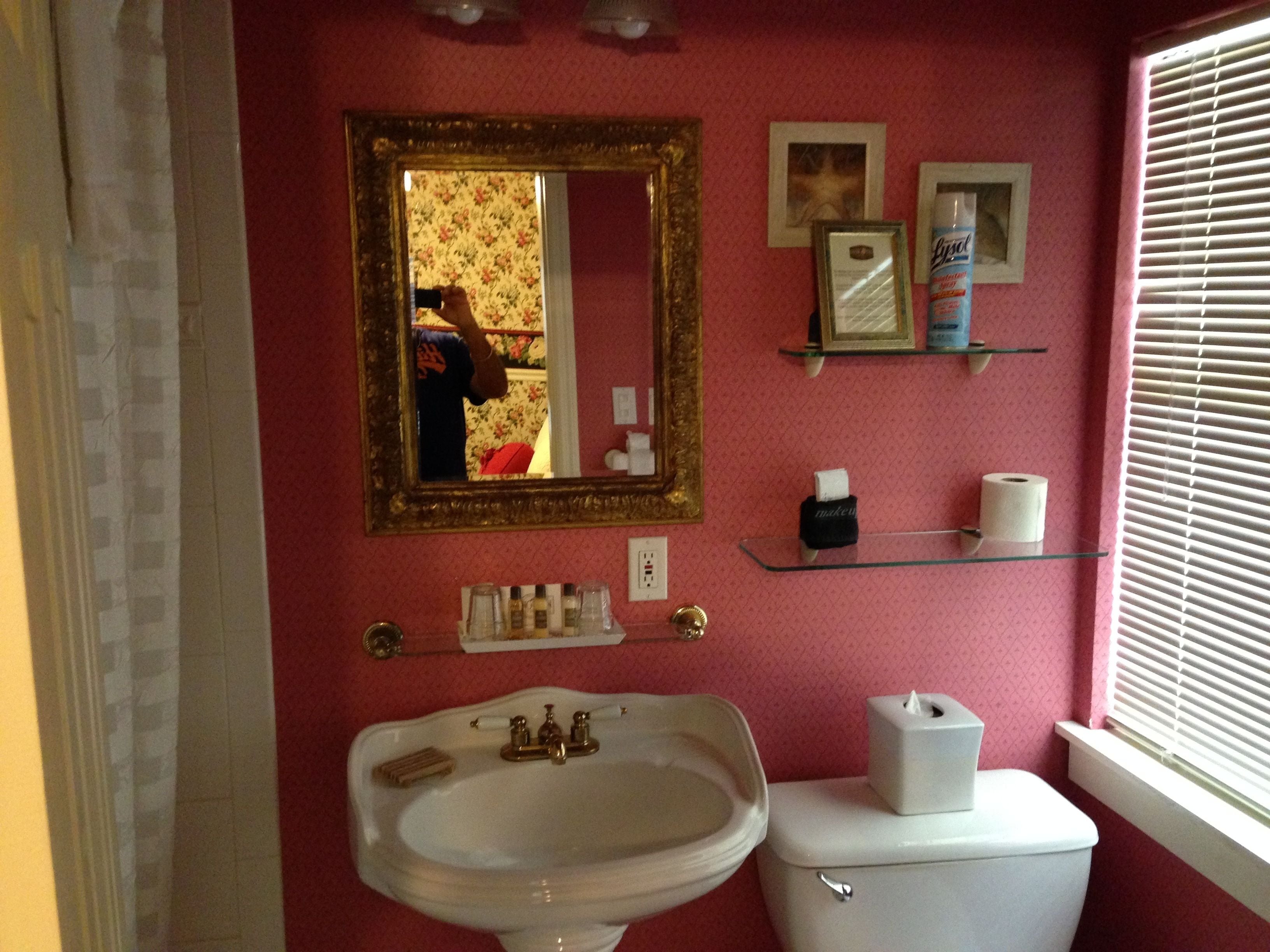 Un baño con lavabo, inodoro y espejo. Hay estantes con artículos de aseo y un cuadro en la pared.