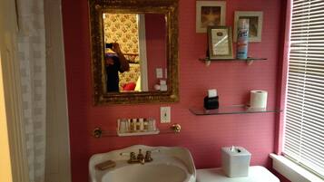 Un baño con lavabo, inodoro y espejo. Hay estantes con artículos de aseo y un cuadro en la pared.