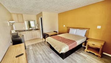 A compact hotel room with a bed, bedside table, a small desk with a chair, a kitchenette, and a bathroom.