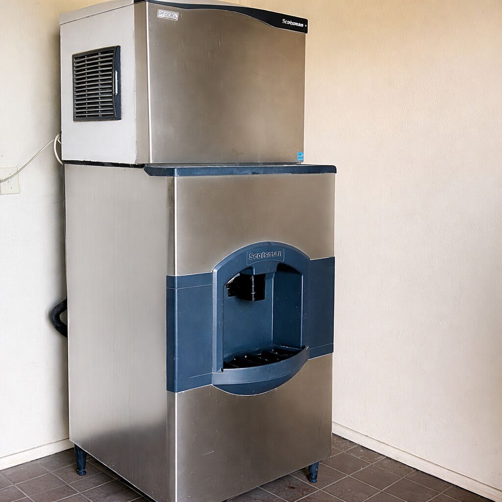 A stainless steel ice and water dispenser with a blue accent, located in a corner against a plain wall.