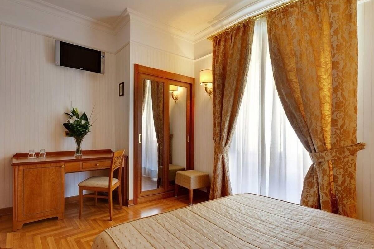 A hotel room with a wooden desk, chair, and bedside table. There is a television mounted on the wall, a mirror, and a window with curtains.