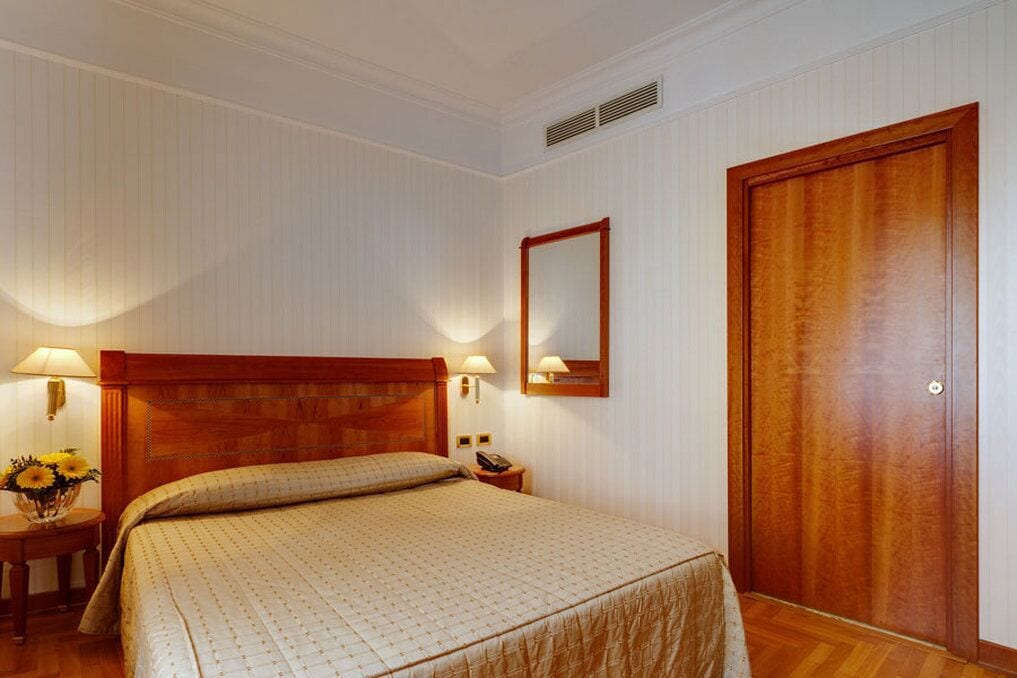 A hotel room with a wooden bed, two bedside lamps, a nightstand with a vase of flowers, a wooden door, and a wall-mounted mirror.
