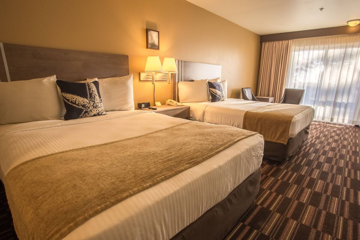 Ein Hotelzimmer mit zwei Betten, einem Nachttisch mit Lampe, einem Sessel und einem großen Fenster mit Vorhängen.