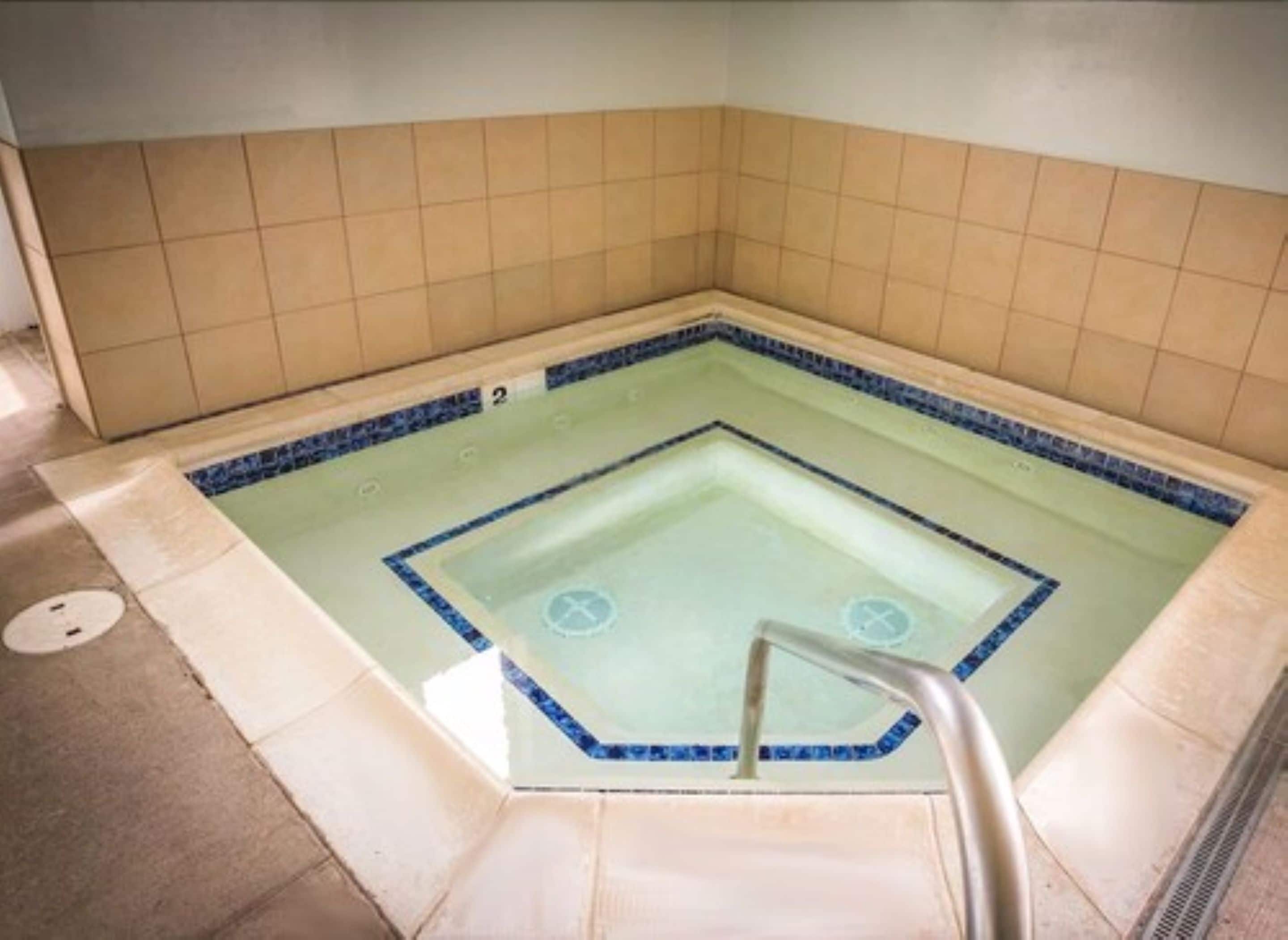 Un jacuzzi carré avec un bordure bleue, des murs carrelés et une échelle en métal.