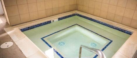 Un jacuzzi carré avec un bordure bleue, des murs carrelés et une échelle en métal.