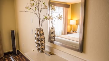 A hotel room with a bed, a desk with a chair, a vase with flowers, and a mirror reflecting the room.