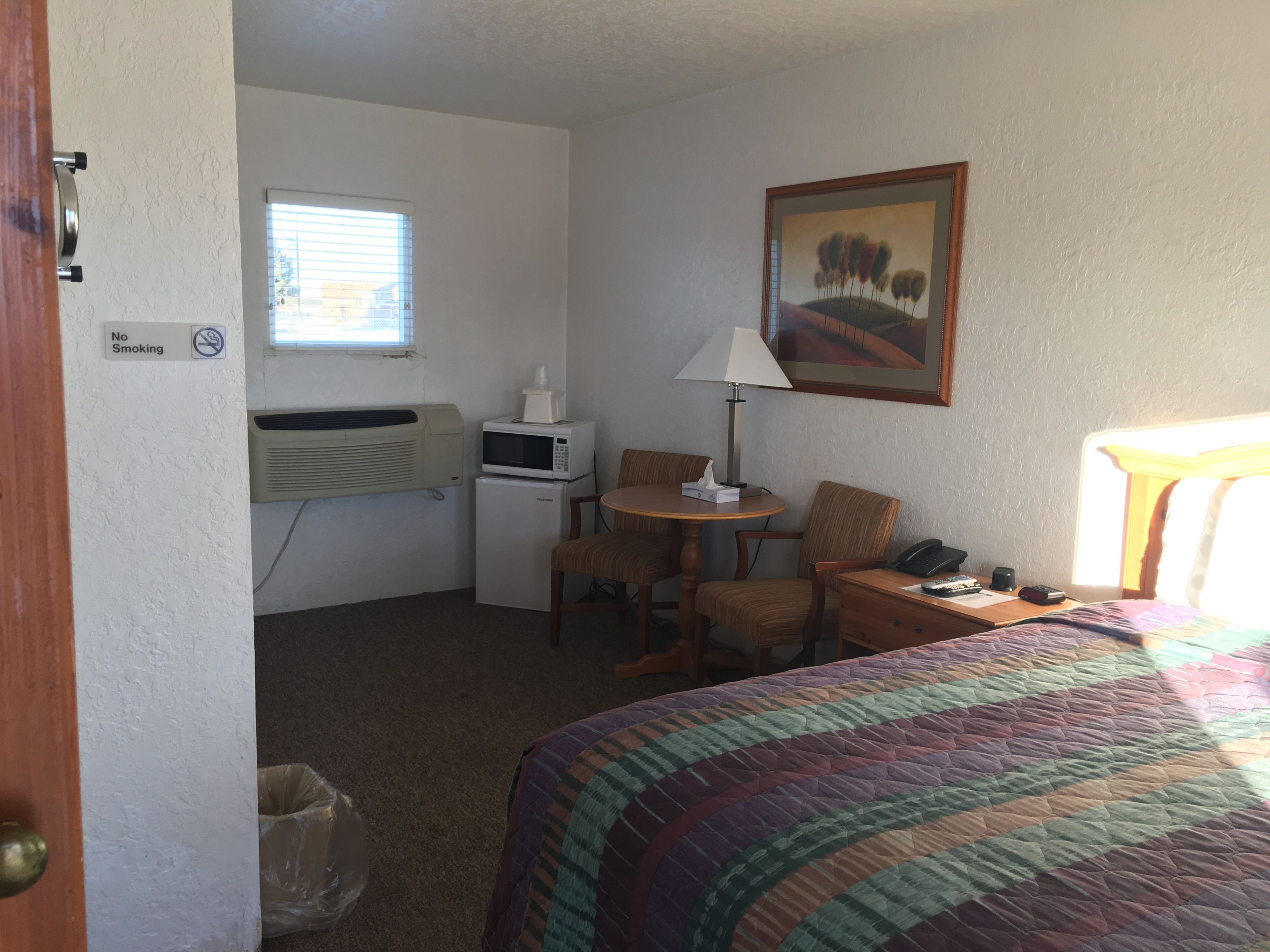 A hotel room with a bed, a desk with a lamp, a microwave, a refrigerator, a chair, a trash bin, and a painting on the wall.
