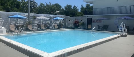 Une piscine rectangulaire d’une profondeur de 1,80 mètre, entourée de chaises longues et de parasols, avec un panneau indiquant « INTERDIT DE PLONGER ».