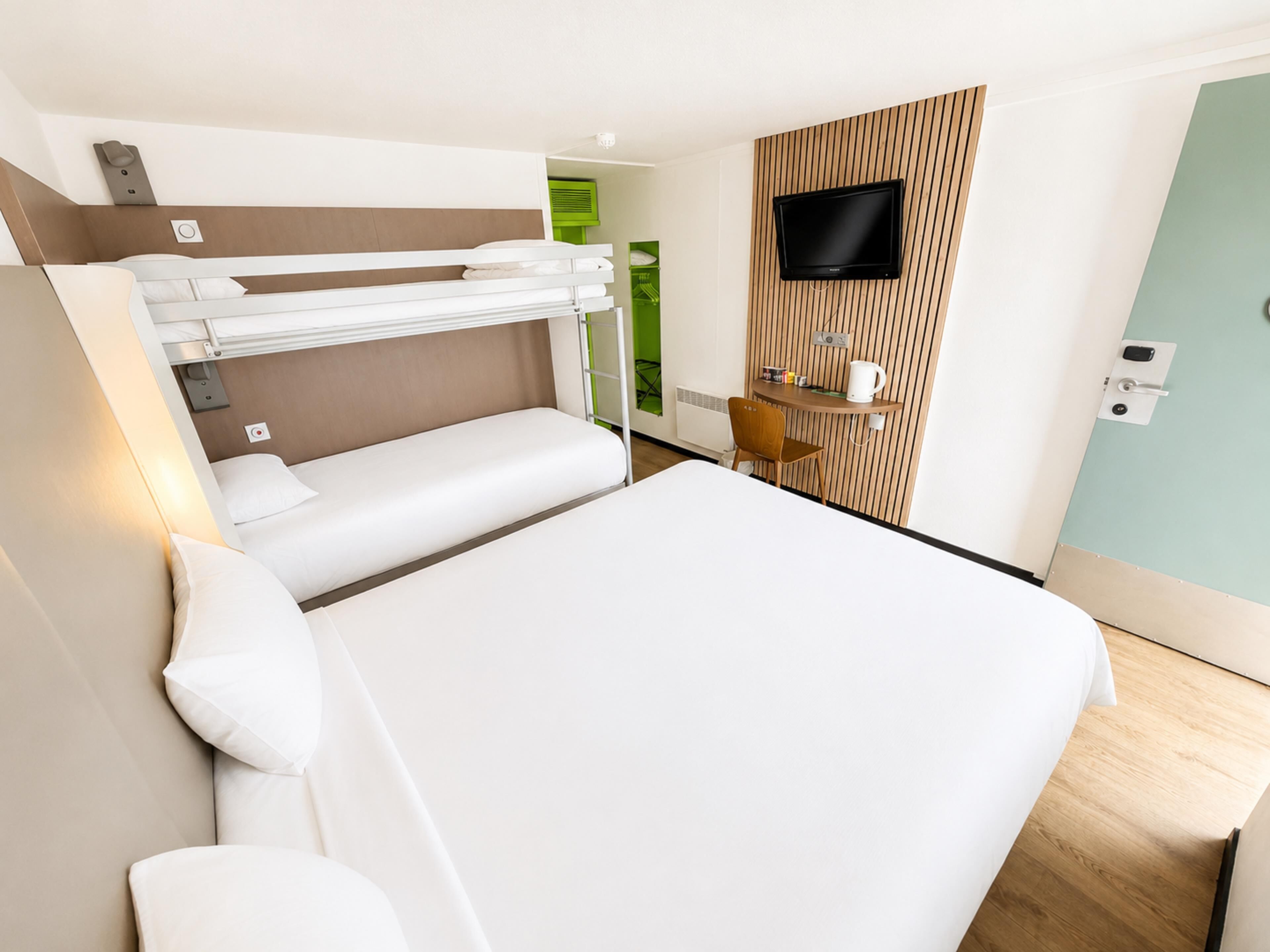 A hotel room with a bunk bed, a desk with a chair, a flat-screen TV mounted on the wall, and a small window.