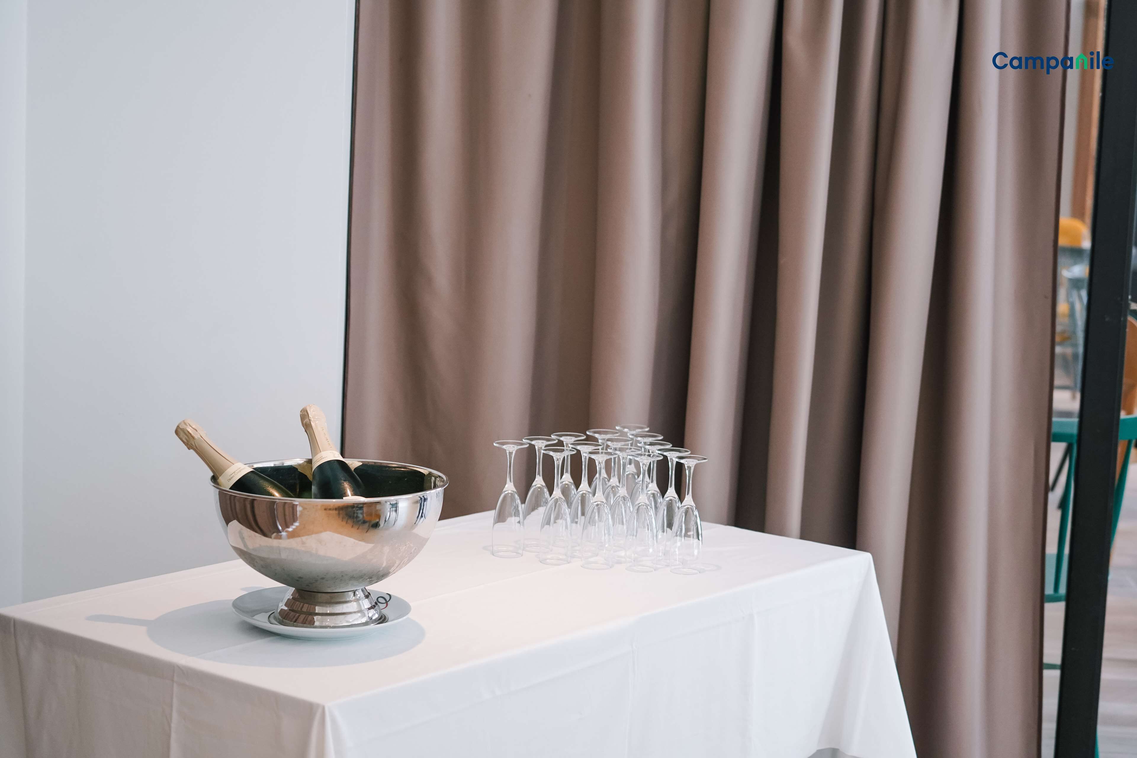 Una mesa con un bol de plata que contiene botellas de champán y copas, frente a un fondo de cortinas.
