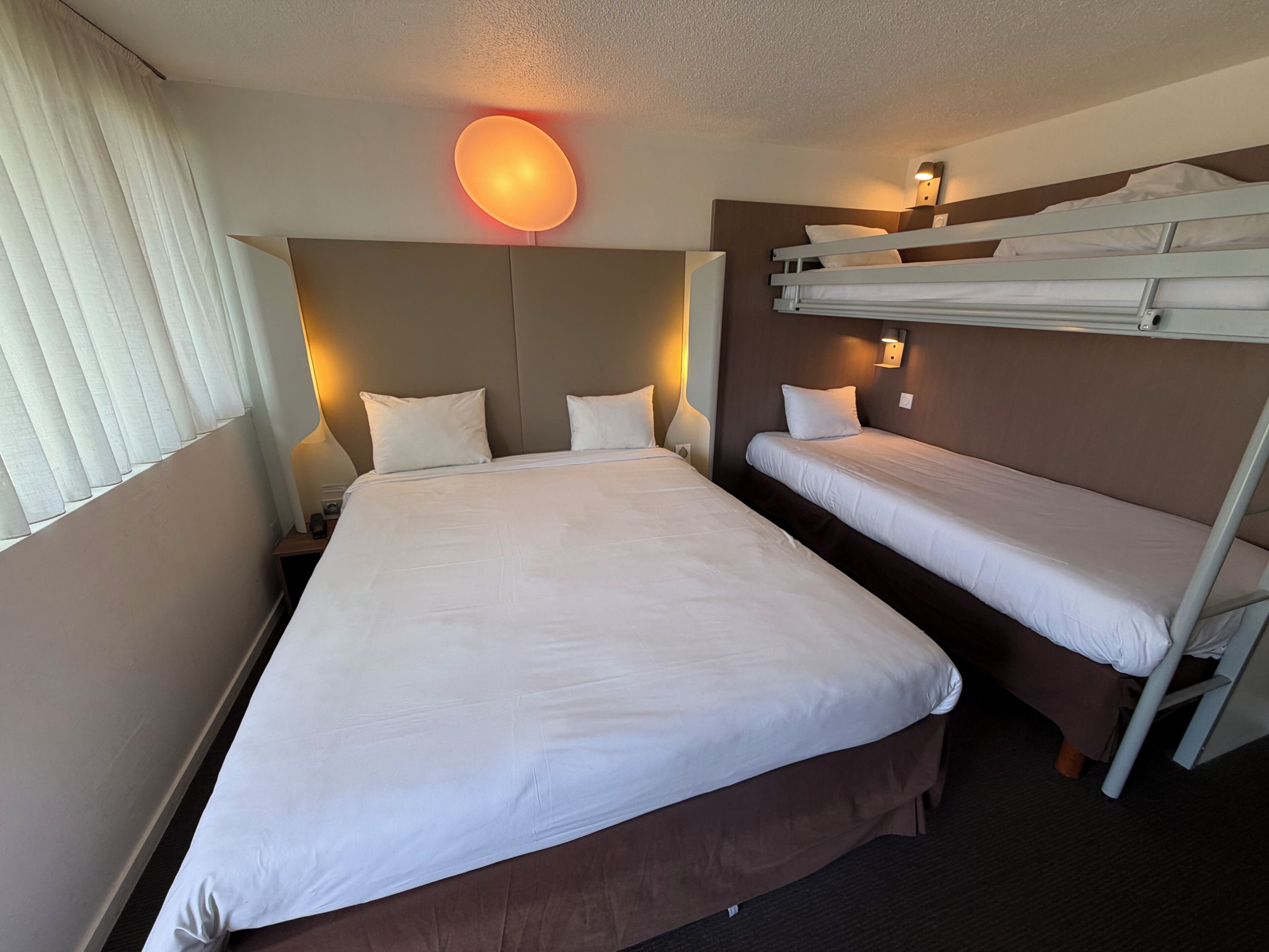 Une chambre d’hôtel avec un grand lit, deux petits lits, une table de chevet, une lampe et un luminaire fixé au mur.