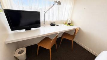 A hotel room with a white desk, a television, a lamp, two wooden chairs, a coffee maker, and a trash bin.
