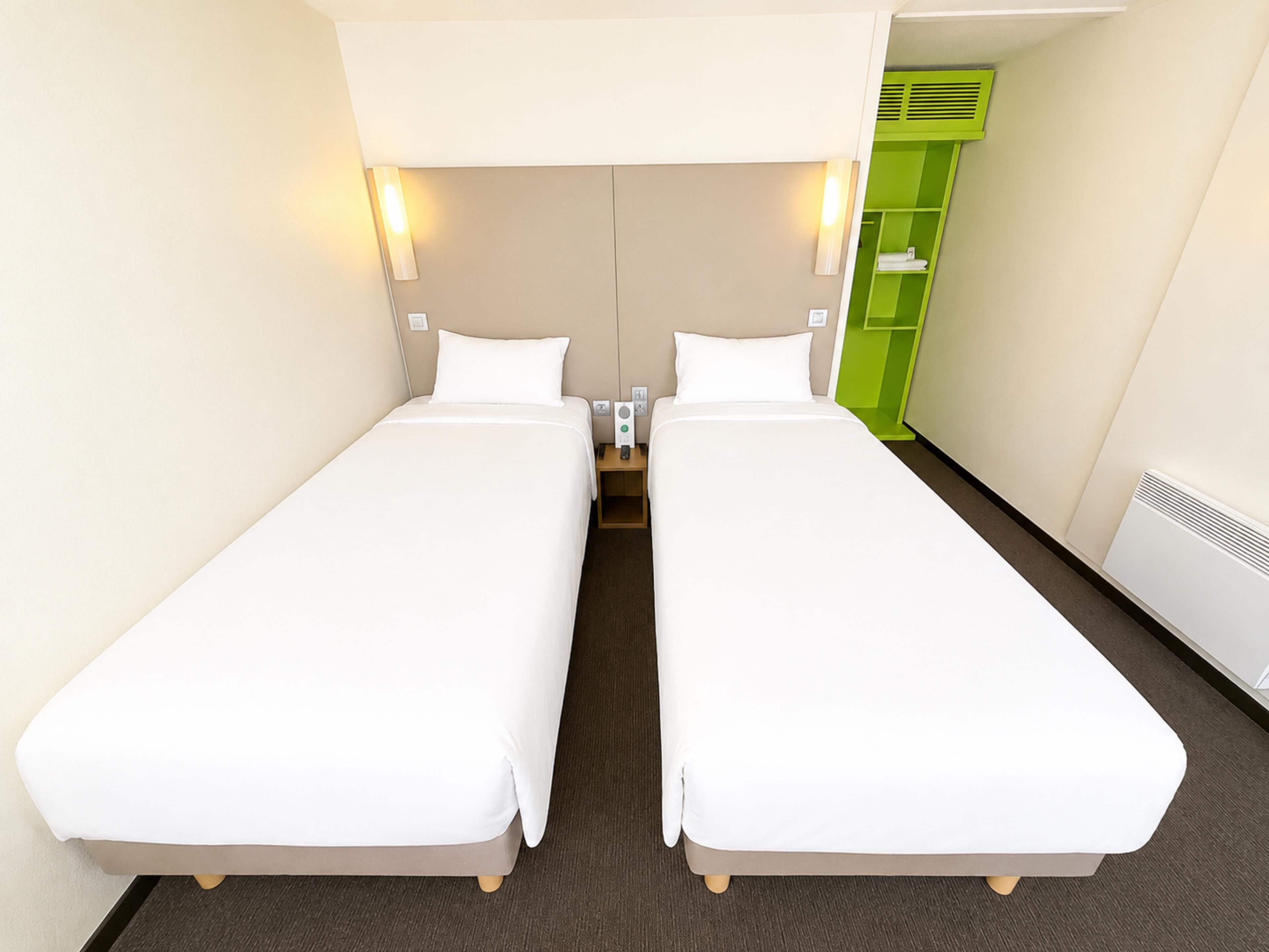 Una habitación de hotel con dos camas individuales, una mesita de noche con lámpara y un mueble de almacenamiento verde.