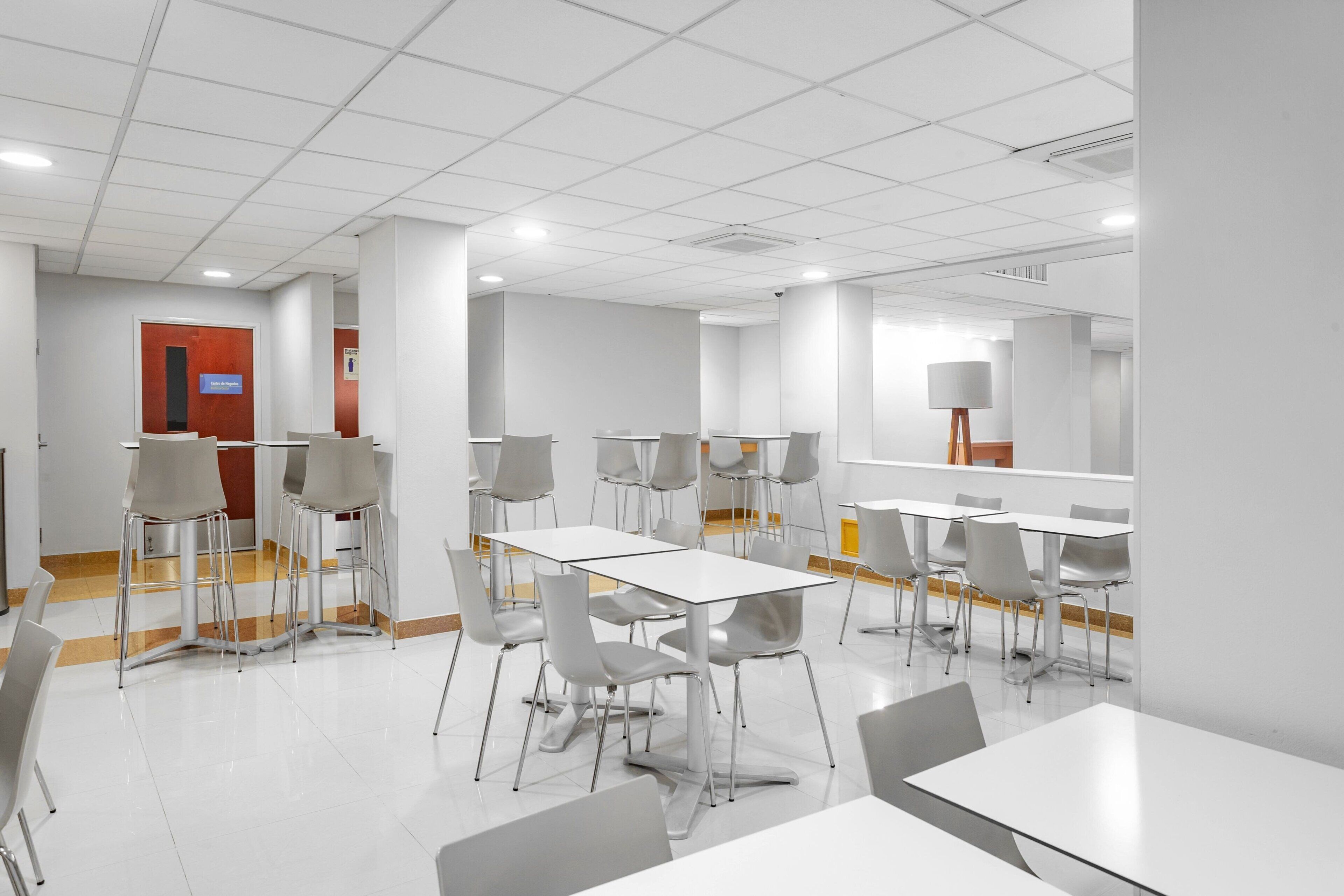 Un espace repas moderne avec des tables et des chaises blanches, une porte rouge et une grande lampe.