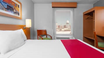 Una habitación de hotel con cama, una silla, un ventanal con vistas, perchero y un cuadro en la pared.