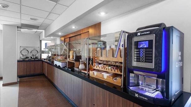A modern coffee station with a Caffenio espresso machine, a glass display case, and a variety of coffee beans and pastries. - City Express by Marriott Celaya Parque (Celaya)