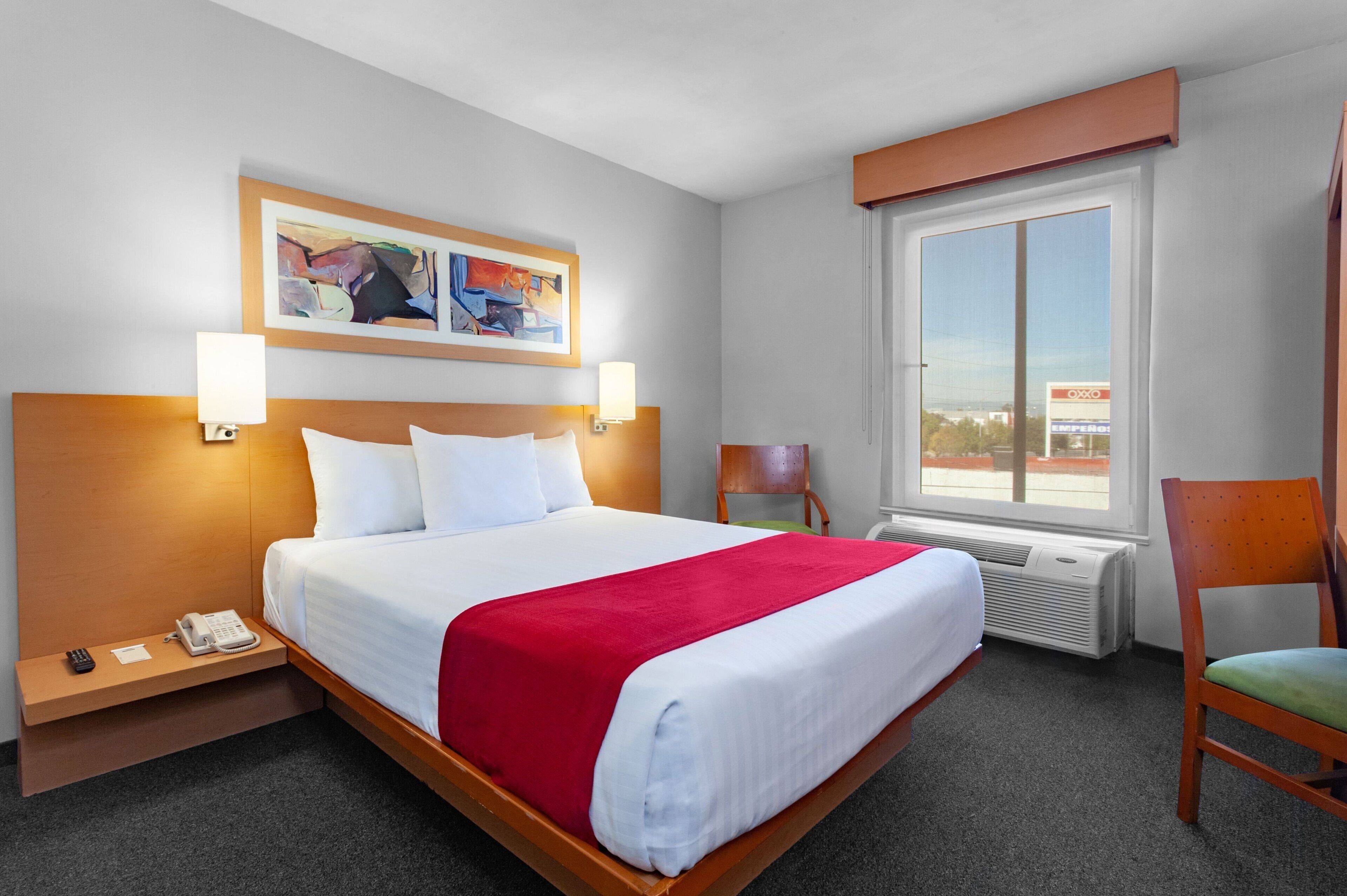 A hotel room with a large bed, a bedside table with a phone, a chair, a window with a view of a gas station, and a painting on the wall.