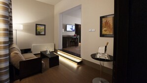 Una habitación de hotel moderna con sofá, una mesa de centro, una mesita auxiliar, una lámpara, un cuadro enmarcado, un televisor y una puerta que da a un comedor.