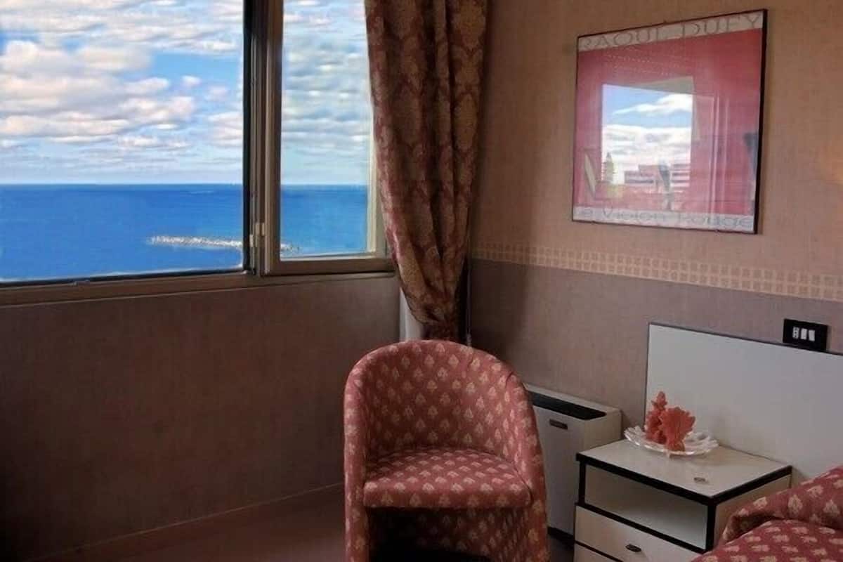A hotel room with a large window offering a view of the sea and clouds. There is a patterned armchair and a small bedside table with a decorative item.