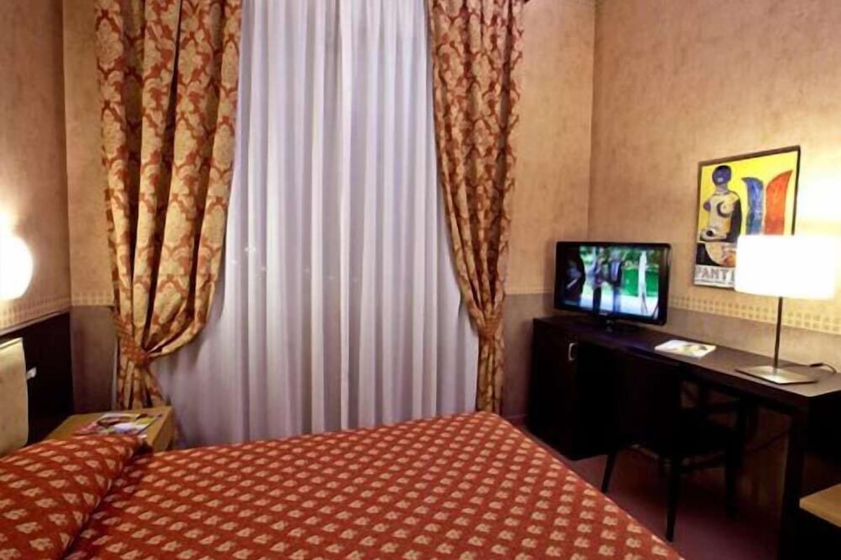 A hotel room with a bed, curtains, a desk with a television, and a lamp.