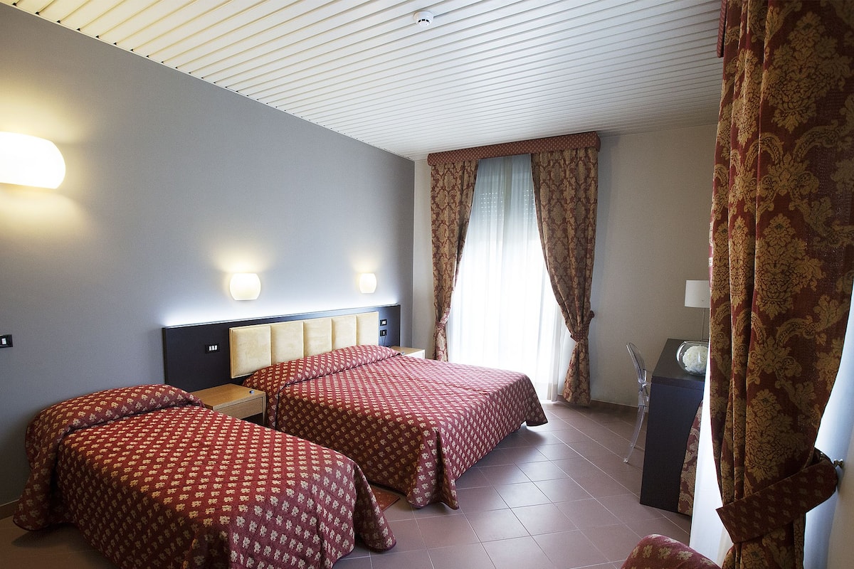 A hotel room with two beds, a nightstand, a lamp, and a window with curtains.
