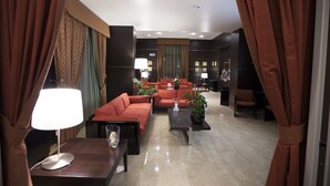 Un moderno vestíbulo de hotel con un área de estar que incluye sofás rojos y una mesa de centro, un televisor sobre un mueble bajo y un mostrador de recepción.