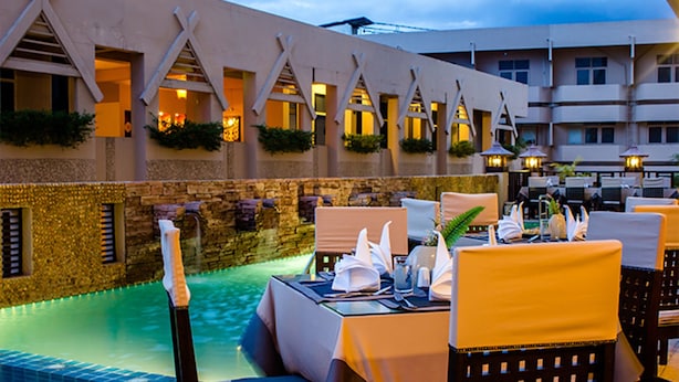 Un espace repas aménagé au bord de la piscine, avec des tables pour deux, un éclairage extérieur et un mur en pierre agrémenté de verdure.