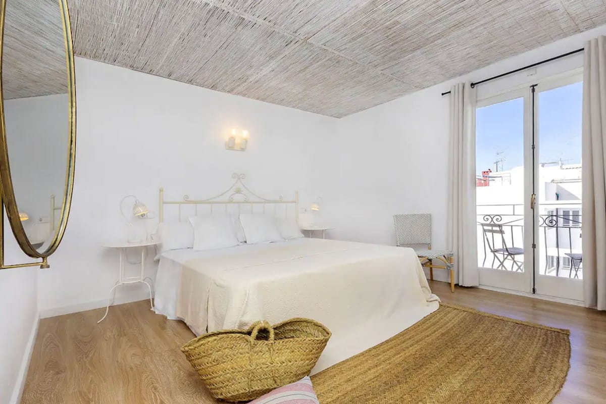 A bedroom with a large bed, a bedside table, a lamp, a mirror, and a view of the outside balcony.