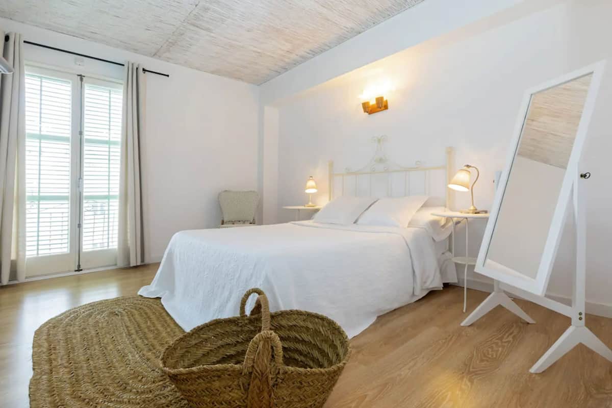A bedroom with a bed, bedside lamps, a standing lamp, a woven basket, and a large window with curtains.