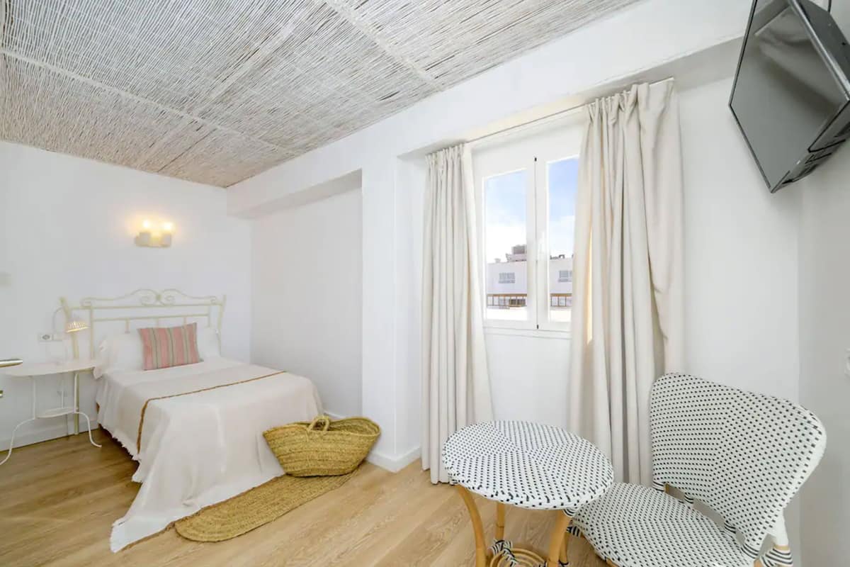 A bedroom with a bed, bedside table, lamp, and a view of the city through a window.