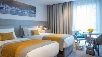 A hotel room with two beds, a desk, a chair, a table with a vase of flowers, and a cityscape painting on the wall.