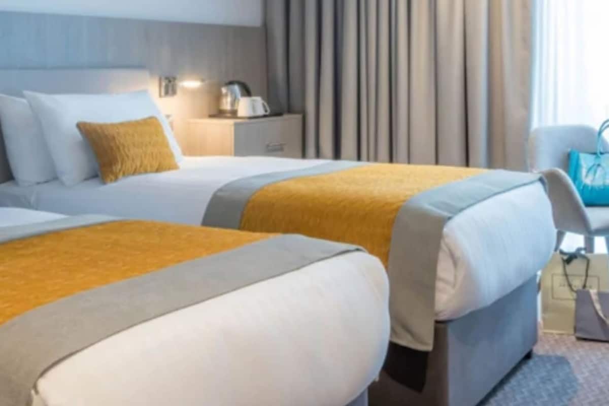 Habitación de hotel con dos camas, una mesita de noche, una lámpara y vistas al exterior.