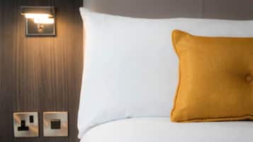 Une chambre d’hôtel avec un lit blanc, un oreiller de couleur moutarde et un luminaire fixé au mur.