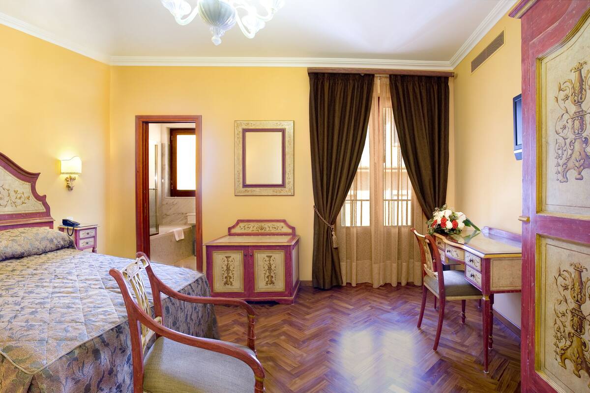 Un dormitorio con una cama grande, una silla de madera, una mesita de noche, un aparador y un escritorio con silla. Hay un espejo en la pared y una ventana con cortinas.