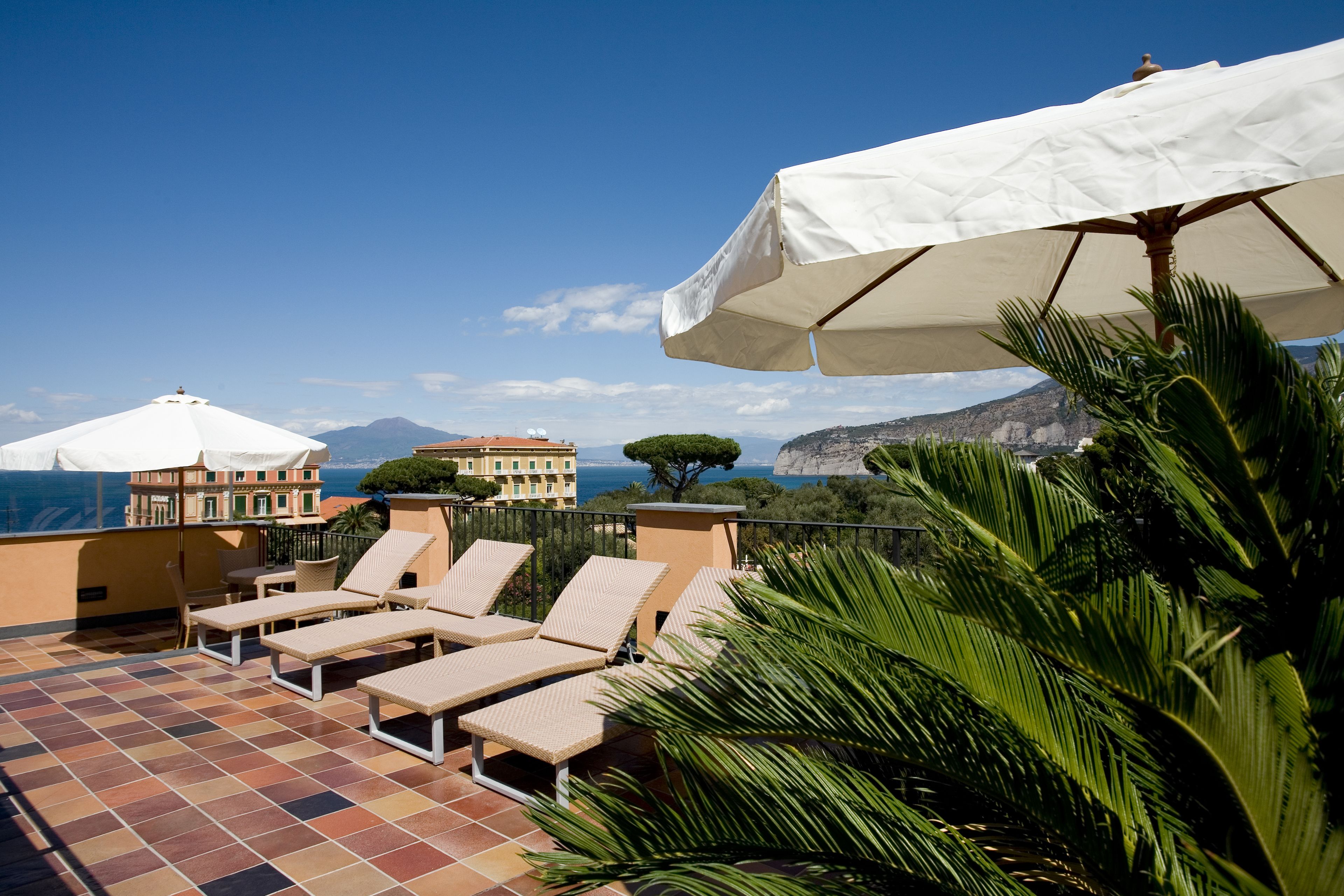 Eine Terrasse mit Liegestühlen und Sonnenschirmen mit Blick auf eine malerische Küstenlandschaft, mit Gebäuden und Bergen.