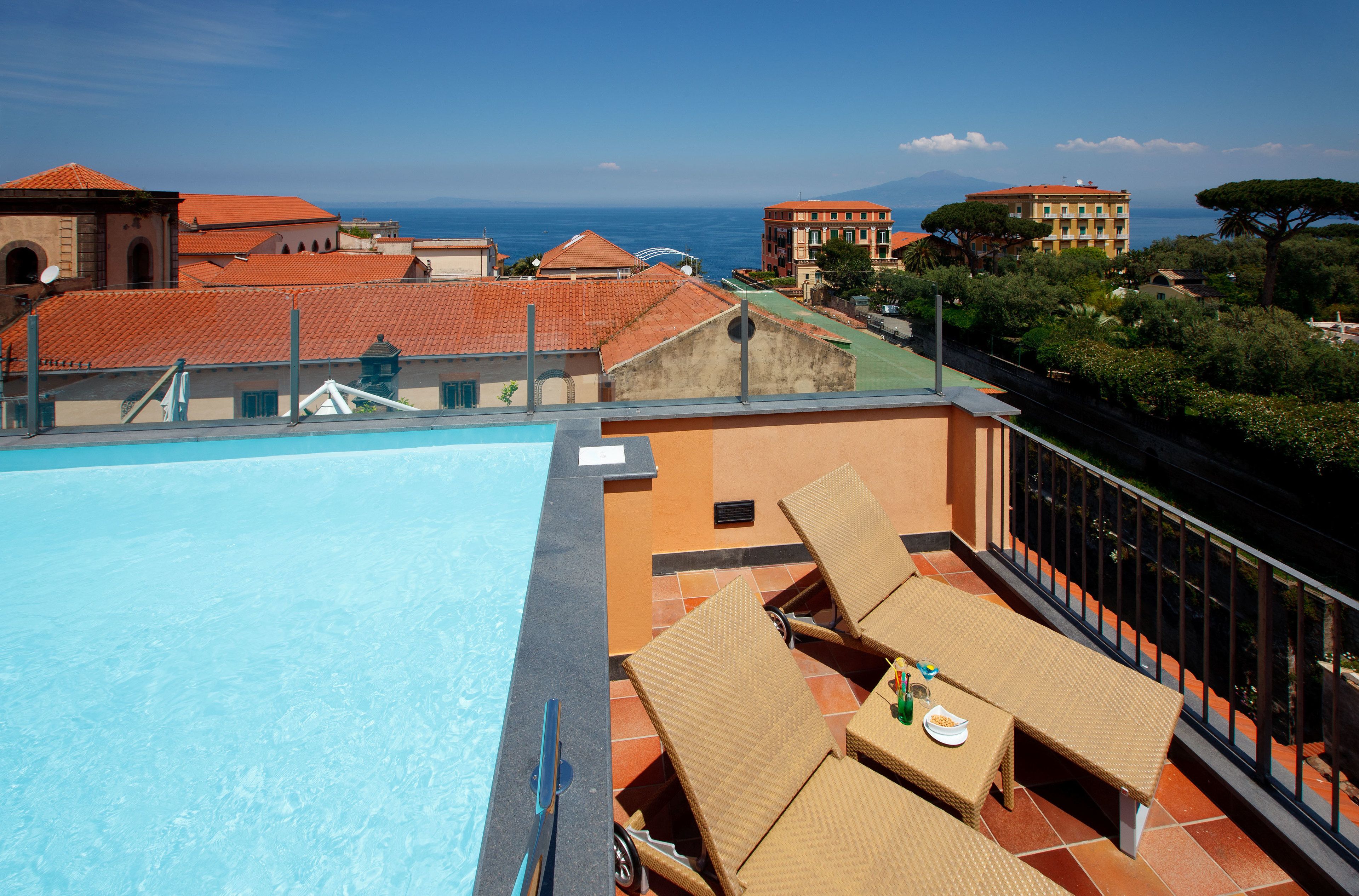 Una piscina en la azotea con sillones de descanso y una mesa, con vistas a un paisaje urbano con edificios de tejados rojos y vistas al mar.